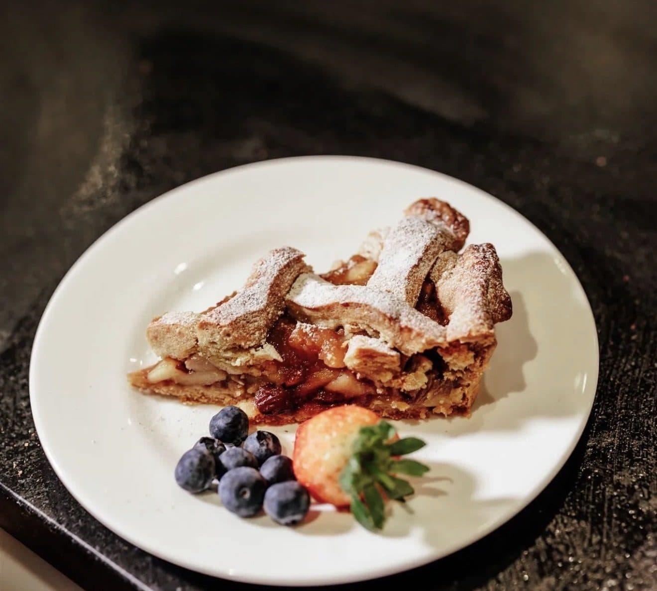 A slice of homemade apple pie dusted with icing sugar, served with blueberries and a strawberry
