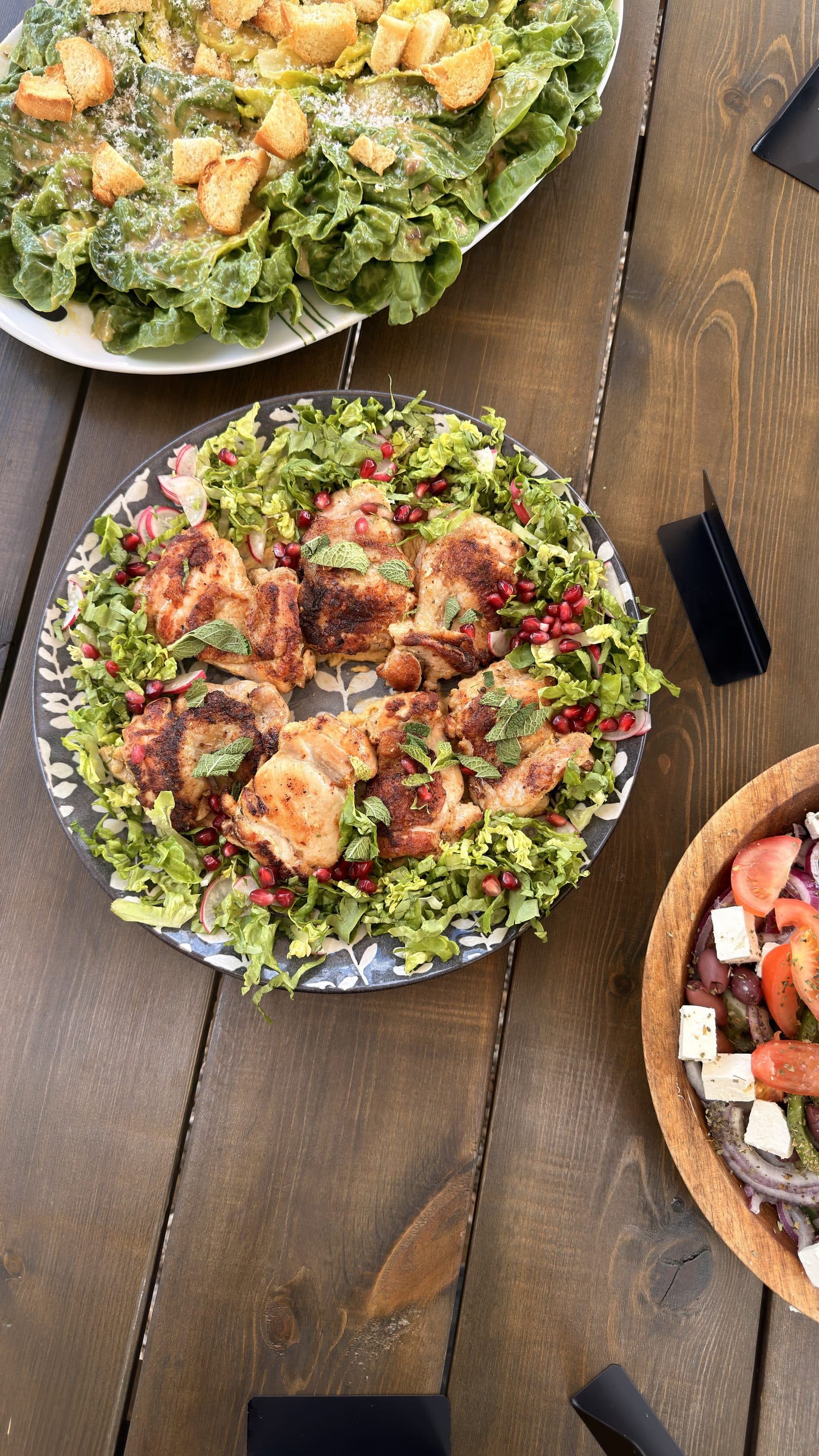 A platter of marinated chicken on a bed of dressed leaves with pomegranate, set on a wooden table with salads alongside