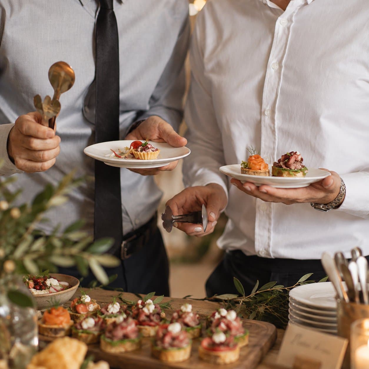 Guests in shirts and ties holding plates of canapés beside a laden serving board