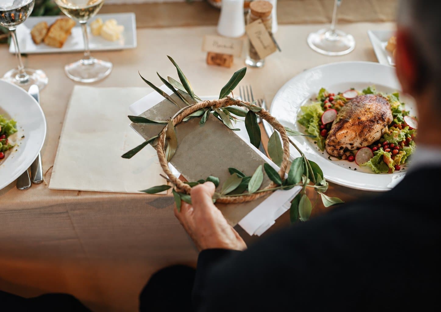 A wedding guest at a laid reception table holding an olive-wreath place card, a plated main beside them