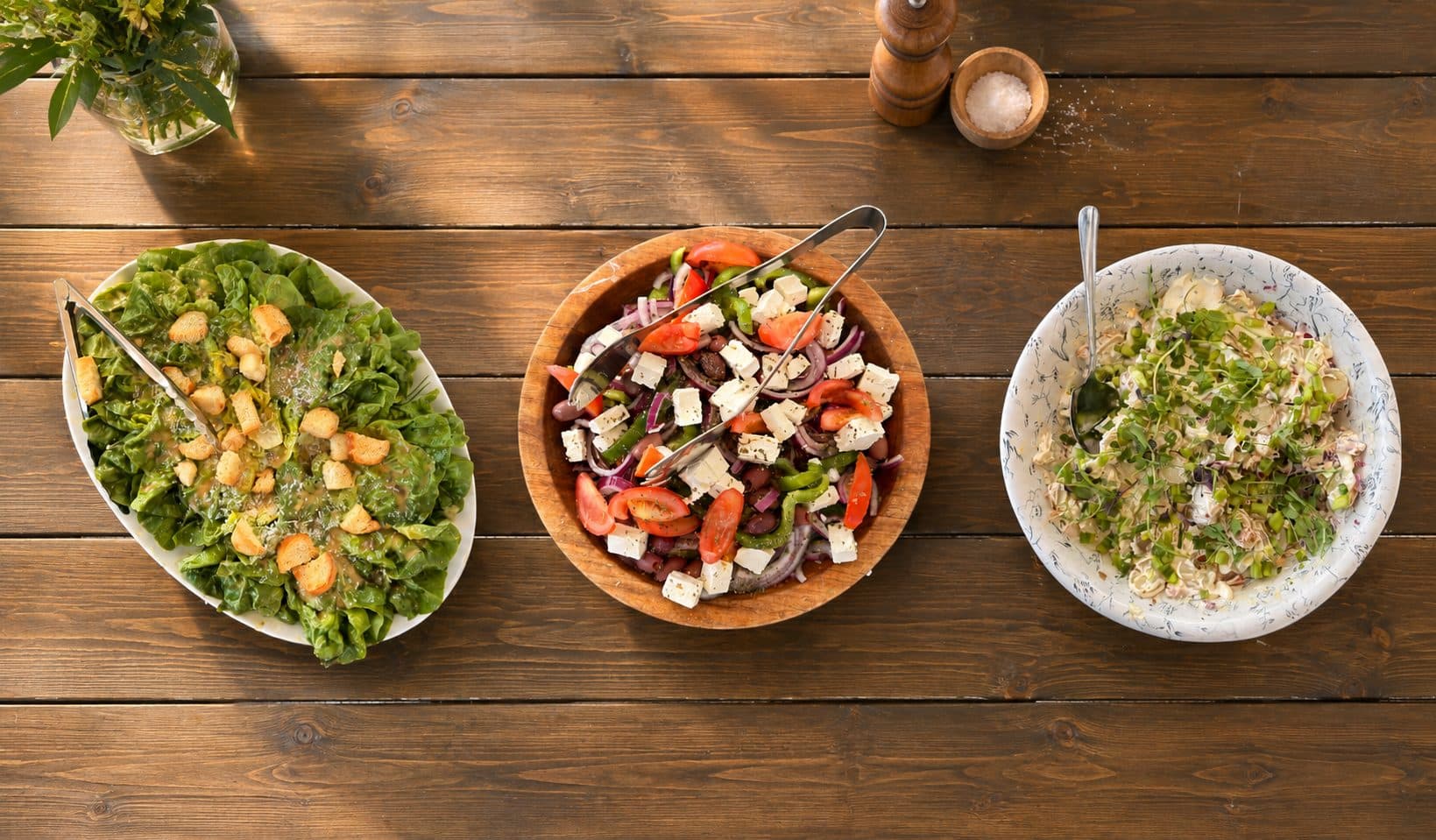 Three sharing bowls of salad laid along a wooden table with serving tongs