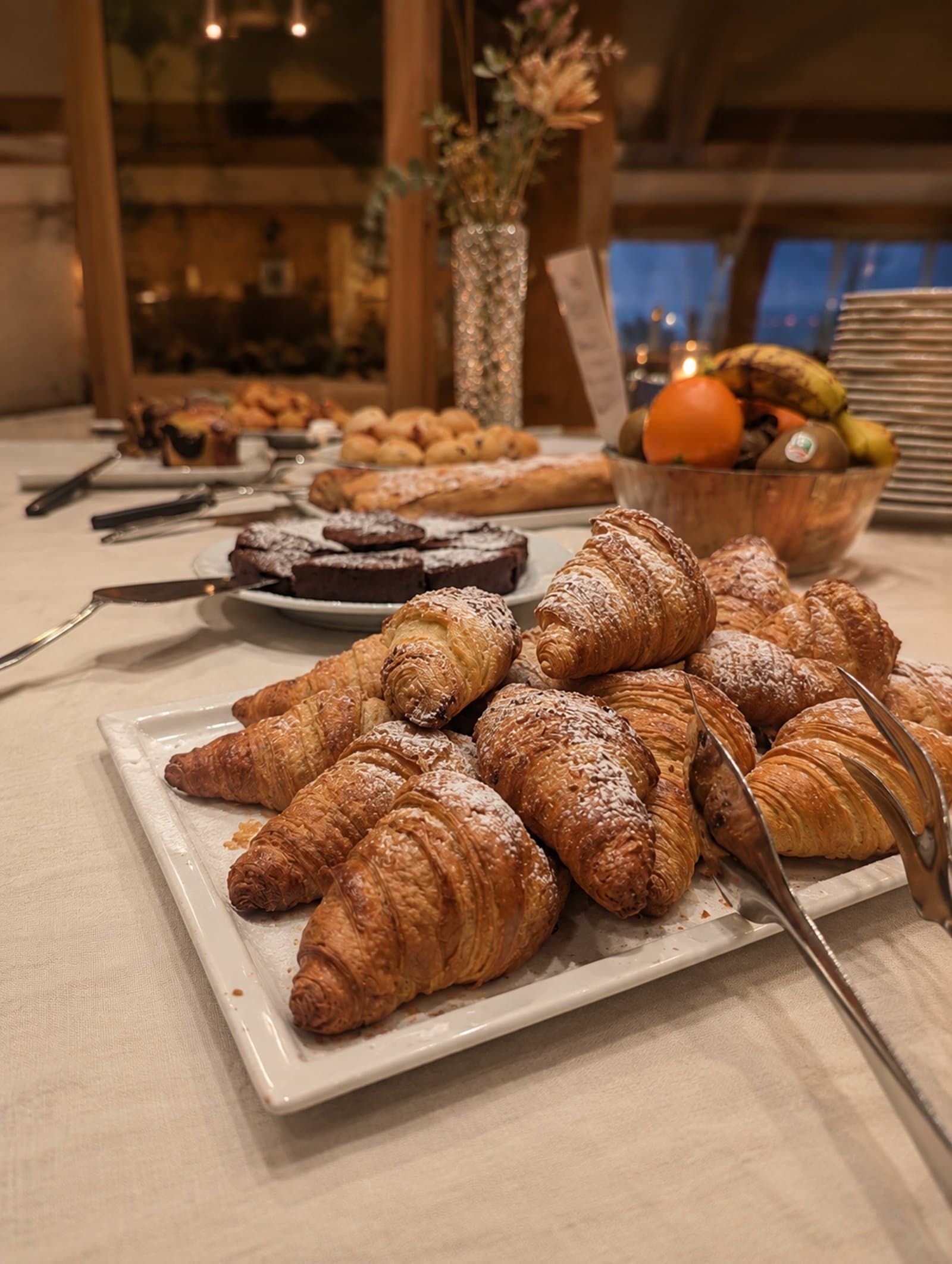 A breakfast buffet platter of croissants dusted with icing sugar, with brownies and fruit behind